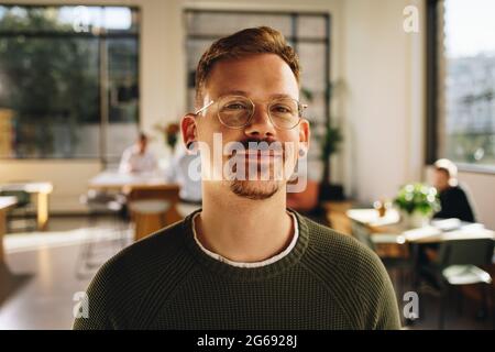 Geschäftsmann lächelt, während er im kooperativen Bereich mit Leuten steht, die hinten arbeiten. Glücklicher Mann, der im Amt steht und Kollegen arbeiten. Stockfoto