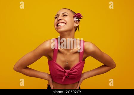 Hübsche junge Frau in Casuals, die wegschaute und lächelte. Schöne Frau trägt Blumen auf rasierten Kopf vor gelbem Hintergrund. Stockfoto