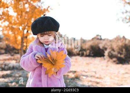 Kind Mädchen 3-4 Jahre alt tragen schwarze Baskenmütze und flauschig rosa Herbst Mantel im Park über gelben Blättern im Freien Nahaufnahme. Herbstsaison. Glück. Stockfoto