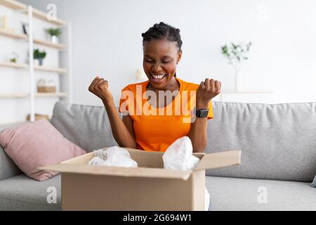 Aufgeregt schwarze junge Frau Auspacken Papppaket im Wohnzimmer, macht JA Geste, lieben ihr bestelltes Produkt Stockfoto