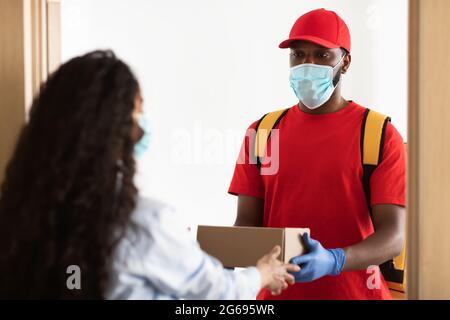 Schwarzer Lieferer in Maske, der eine Kiste an weibliche Kundin gibt Stockfoto