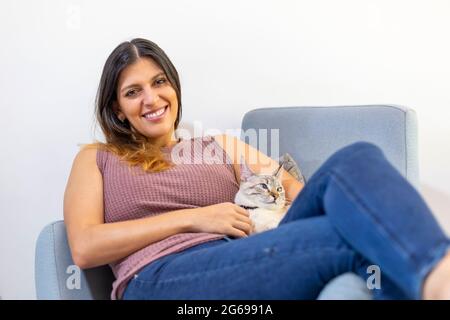 Porträt einer jungen Latina-Frau, die mit ihrer Katze auf einem blauen Stuhl liegt Stockfoto