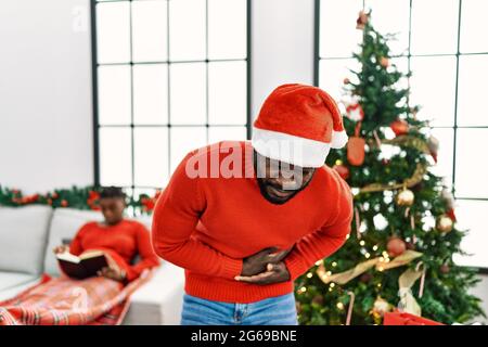 Junger afroamerikanischer Mann, der am weihnachtsbaum mit der Hand auf dem Bauch steht, weil Übelkeit, schmerzhafte Krankheit, Unwohlsein. Schmerzkonzept. Stockfoto