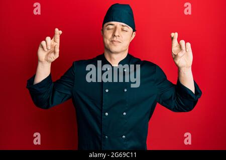Schöner junger Mann in professioneller Koch-Uniform und Hut gestikuliert Finger überquert lächelnd mit Hoffnung und Augen geschlossen. Glück und abergläubischen concep Stockfoto