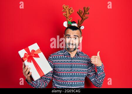 Junger hispanischer Mann trägt Hirsch weihnachtshut mit Geschenk zeigt Daumen nach oben auf die Seite lächelnd glücklich mit offenem Mund Stockfoto