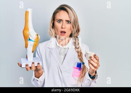 Schöne junge blonde Ärztin hält anatomisches Modell des Kniegelenks und Proteinpuders im Schockgesicht und sieht skeptisch und sarkastisch aus, Überraschung Stockfoto