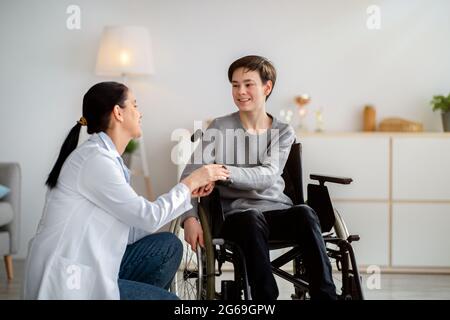 Unterstützung der Gesundheitsfürsorge für behinderte Menschen. Arzt hält während eines Arztbesuchs die Hände eines behinderten Teenagers im Rollstuhl Stockfoto