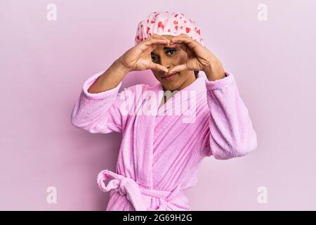 Hispanic Mann trägt Make-up mit Duschhandtuchkappe und Bademantel tun Herzform mit Hand und Finger lächelnd Blick durch Zeichen Stockfoto