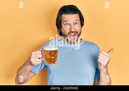 Kaukasischer Mann mittleren Alters, der ein Glas Bier trinkt und den Daumen zur Seite zeigt und glücklich mit offenem Mund lächelt Stockfoto