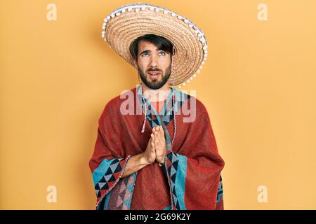 Junger hispanischer Mann mit mexikanischem Hut betteln und beten mit Händen zusammen mit Hoffnungsausdruck im Gesicht sehr emotional und besorgt. Betteln. Stockfoto
