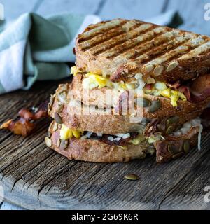 Zwei gegrillte Frühstückspaninis mit Ei und Speck auf einem rustikalen Holzbrett. Stockfoto