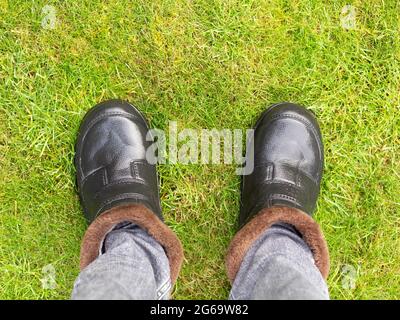 Blick hinunter auf schwarze Gummistiefel mit Fell innen auf regnerisch nassem grünem Gras Stockfoto