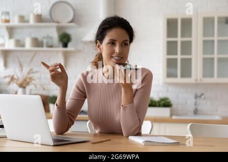 Lächelnde junge Frau aus den 30er Jahren, die eine Audiobotschaft aufzeichnet und am Computer arbeitet. Stockfoto