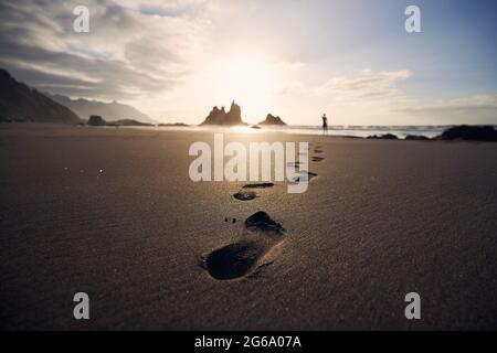 Fußabdrücke im Sand gegen die Silhouette der Person. Mann, der bei goldenem Sonnenuntergang am Strand entlang zum Meer läuft. Teneriffa, Kanarische Inseln, Spanien. Stockfoto