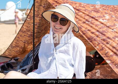 Eine Frau in einem Hut und einem weißen Hemd von der Sonne sitzt in der Nähe eines Touristenzeltes an der Küste. Reisen und Tourismus. Stockfoto