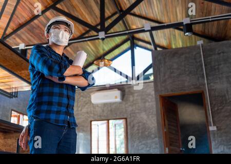 Glücklicher asiatischer Bauingenieur-Techniker, der mit gekreuzten Armen steht, nachdem er die Holzstruktur unter dem Dach bei Construction si überprüft hat Stockfoto