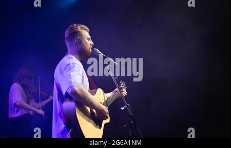 Ein junger Mann in einem weißen T-Shirt mit Bart hält eine akustische Gitarre in der Hand und singt in ein Mikrofon, das auf der Bühne steht Stockfoto