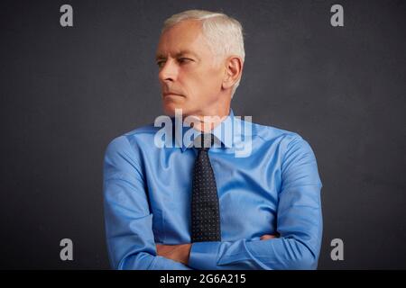 7/8 Schuß von Stattlichen älteren Mann mit Hemd und Krawatte beim Stehen mit verschränkten Armen an dunklen grauen Hintergrund. Stockfoto