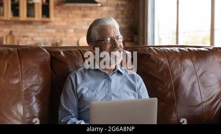 Glücklicher, reifer Mann aus den 70ern, der einen Laptop benutzt und auf dem Sofa sitzt Stockfoto