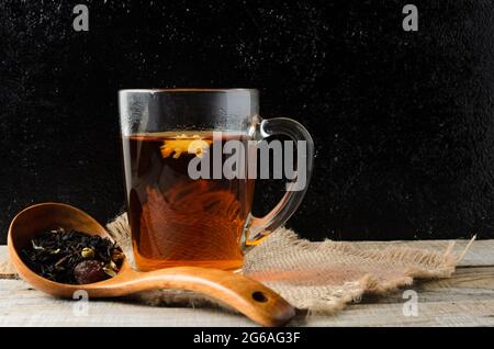 Ein Glas mit duftendem, gebrühtem Blumentee auf einem Holztisch mit einem Holzlöffel. Schwarzer Hintergrund. Selektiver Fokus. Stockfoto