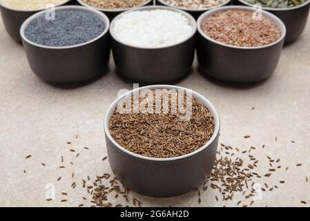 Kümmel. Becher mit Kümmel auf einem Hintergrund verschiedener Samen in schwarzen Behältern auf einer Marmorplatte. Stockfoto