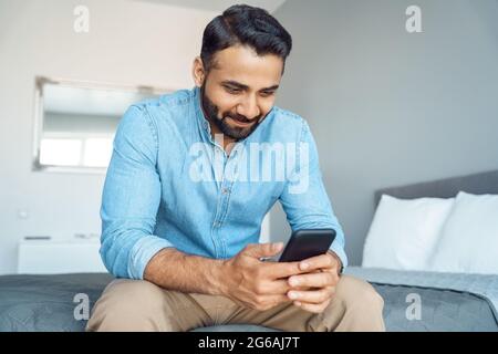 Indischer Mann, der E-Mail-Nachrichten abcheckt und über ein soziales Netzwerk telefoniert, sitzt auf dem Bett Stockfoto