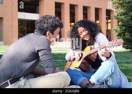 Ein junger lateinmann lehrt einer jungen lateinerin, die auf dem Universitätscampus lachend Gitarre spielt. Universitätsleben, tausendjährige Generation. Stockfoto