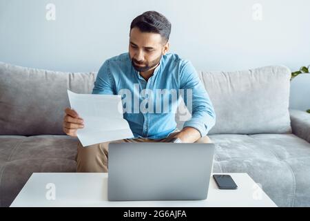 Fokussierter ernster indischer Mann beim Lesen eines Papierbriefes vor dem Laptop Stockfoto