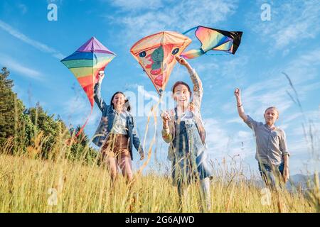 Lächelnde gils und Bruder Junge laufen mit fliegenden bunten Drachen auf der hohen Graswiese in den Bergfeldern. Glückliche Kindheitsmomente oder Outdoor-Ti Stockfoto