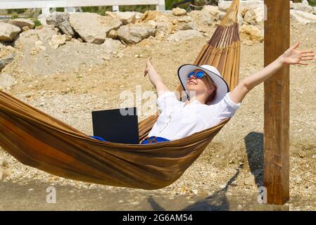 Arbeiten Sie von überall aus. Junge lächelnde Frau, freiberufliche Mitarbeiterin im Strohhut, die am Strand am Laptop arbeitet. Stockfoto
