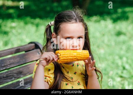 Ein Kind ist eine Ähre aus Mais. Gelber Mais und ein Kind in einem gelben Kleid im Park auf einem Spaziergang. Kindervegetarismus und gesunde Desserts Stockfoto