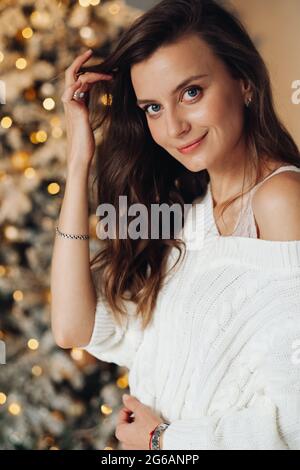 Schöne Dame in weißem Pullover posiert neben geschmückten Weihnachtsbaum Stockfoto