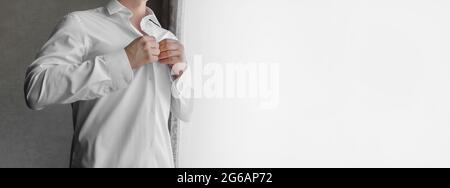 Ein Mann klammelt Knöpfe auf einem weißen Hemd mit seinen Händen im Licht vom Fenster. Stockfoto