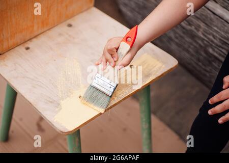 Nahaufnahme der Hand eines Kindes, das eine Bürste hält und einen Holzstuhl zur Wiederverwendung malt Stockfoto