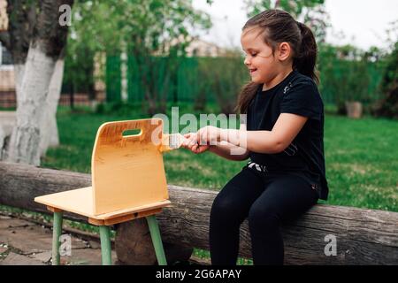 Ein fünfjähriges Mädchen malt einen alten kleinen Stuhl mit gelber Farbe zur Wiederverwendung, während es auf einem Holzbuch im Garten sitzt Stockfoto