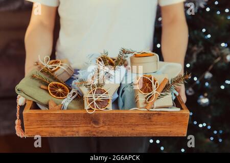 Mann hält ein Holztablett mit umweltfreundlichen eingepackten Weihnachtsgeschenkboxen, die mit natürlichen getrockneten Orangen, Zimtstangen und Fichtenzweig dekoriert sind. Christus Stockfoto