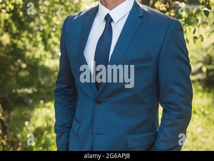 Ein Geschäftsmann in einem weißen Hemd, einem blauen Anzug und einer Krawatte steht mit den Händen in den Taschen draußen im Park. Stockfoto