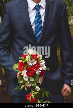 Der Bräutigam in einem weißen Hemd, einem blauen Anzug und einer Krawatte hält den Brautstrauß der Braut aus weißen und roten Rosen-Blüten. Stockfoto