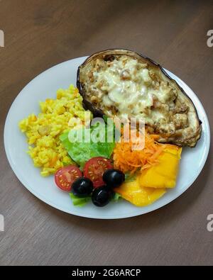 Geröstete Auberginen gefüllt mit Tofu, Pilzen und veganem Käse mit gemischtem Salat und Kurkuma-Reis auf der Seite in weißer Schale. Hausgemachtes Essen im Mittelpunkt Stockfoto