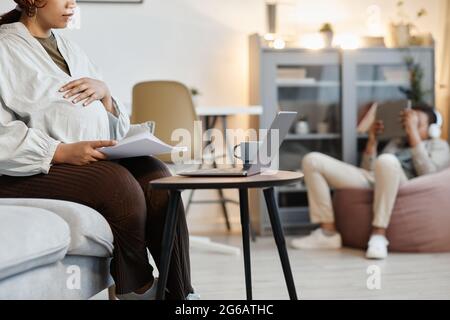 Beschnittenes Porträt einer schwanger afroamerikanischen Frau, die zu Hause einen Laptop benutzt, mit einem Teenager-Sohn im Hintergrund, Kopierbereich Stockfoto