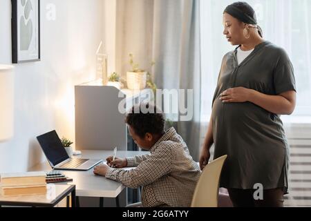 Seitenansicht Porträt einer trächtigen afroamerikanischen Frau, die einem Teenager-Sohn bei Hausaufgaben zusieht, Platz kopieren Stockfoto