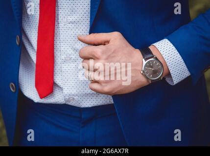 Die Hand eines Mannes mit einer Handuhr hält sich an einem blauen Anzug vor dem Hintergrund eines weißen Hemdes und einer roten Krawatte, einem Kleidungsstil für Geschäftsleute Stockfoto