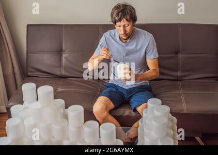 Der Mann isst Joghurt und ist von vielen Joghurtgläsern umgeben, die er in einem Jahr verwendet. Eine riesige Menge von Plastikbehältern für Lebensmittel. Übermäßiger Verbrauch von Stockfoto