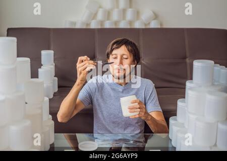 Der Mann isst Joghurt und ist von vielen Joghurtgläsern umgeben, die er in einem Jahr verwendet. Eine riesige Menge von Plastikbehältern für Lebensmittel. Übermäßiger Verbrauch von Stockfoto