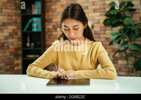Fokussierte junge asiatische Frau, die zu Hause mit einem digitalen Tablet auf dem Schreibtisch sitzt, um in den sozialen Medien zu surfen und nachts zu Hause online zu shoppen Stockfoto