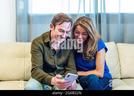 Lächelndes reifes Paar zu Hause, das sich auf dem Handy etwas Lustiges ansieht. Stockfoto