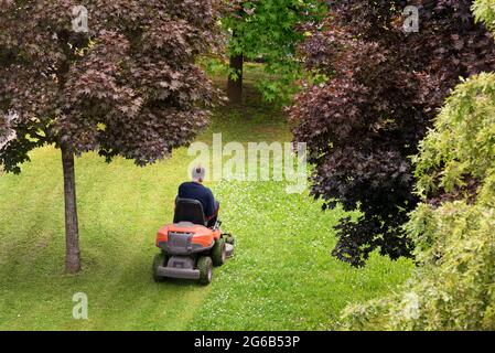 Im Frühling oder Sommer wird ein Mann mit einem Aufsitzmäher in seinem Hinterhof das Gras schneiden, das zwischen grünen Bäumen gesäumt wird Stockfoto