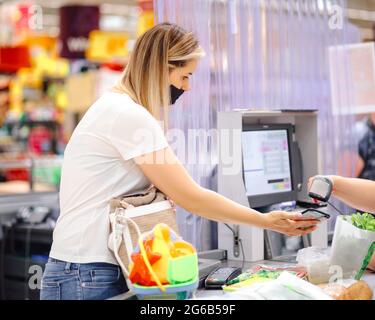 Junge Kundin in individueller Schutzmaske an der Kasse im Supermarkt wird per Handy berechnet. Sicherheitsmaßnahmen und Einkaufen. Moderne Technik Stockfoto