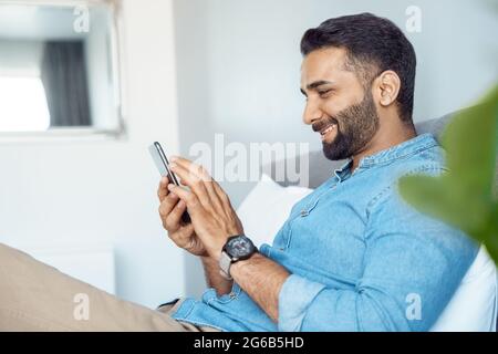Profilporträt eines lächelnden jungen erwachsenen indischen Mannes mit Mobiltelefon-Ruhepause auf dem Bett Stockfoto