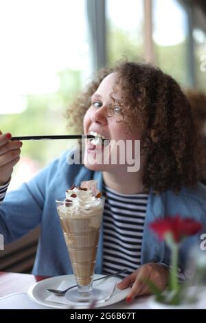 Fröhliche junge, lockige Frau, die im Café am Tisch sitzt und köstlichen Milchshake mit Schlagsahne und Beeren genießt Stockfoto